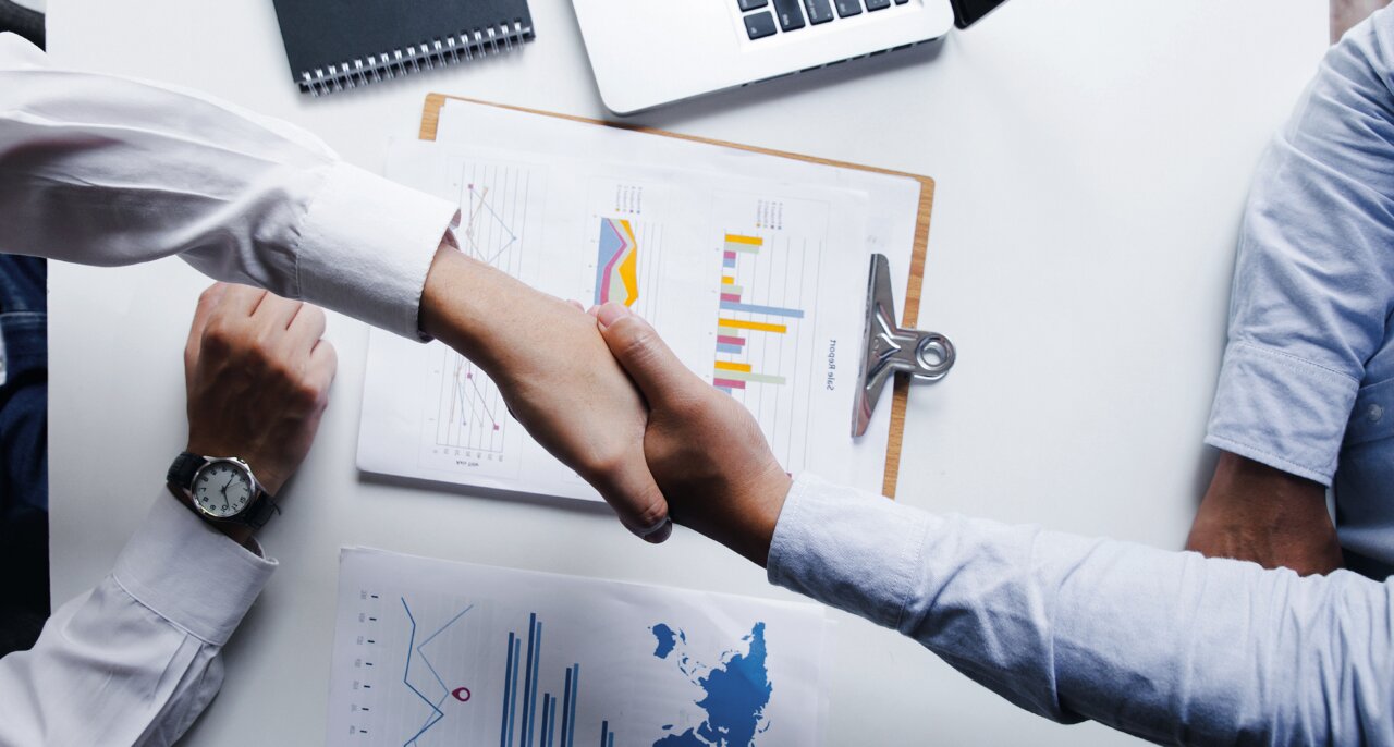 Dos personas estrechando manos sobre una mesa con documentos y gráficos